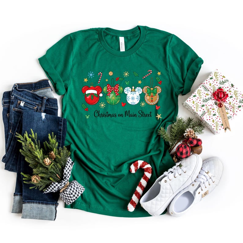 May include: A green t-shirt with a festive design featuring Mickey Mouse-shaped ornaments. The ornaments are red, green, and white, with the text "Christmas on Main Street". The shirt is paired with blue jeans, white trainers, and a wrapped gift.