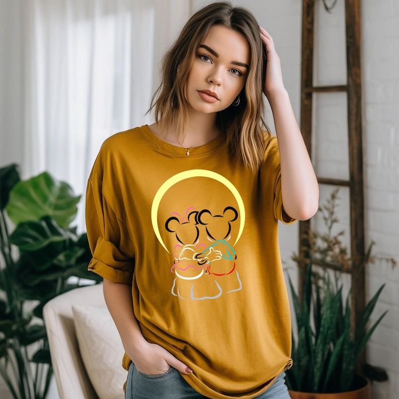 Puede incluir: Camiseta de color amarillo mostaza con una ilustraci&oacute;n de dibujos animados de dos figuras con forma de oso abraz&aacute;ndose dentro de un c&iacute;rculo amarillo. Las figuras tienen contornos negros con acentos rosas, verdes y rojos.