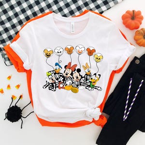 May include: White t-shirt with a graphic of Mickey Mouse, Minnie Mouse, Donald Duck, Goofy, and Pluto dressed as skeletons holding balloons with Halloween-themed designs.