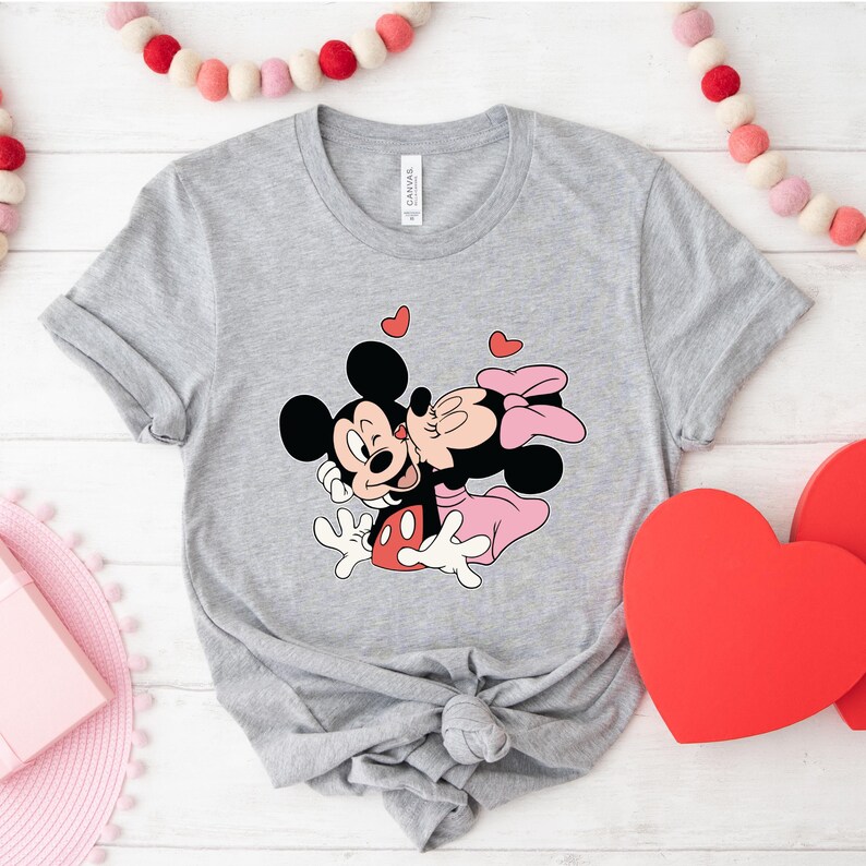 May include: A light gray t-shirt featuring a graphic of Mickey Mouse kissing Minnie Mouse, with red hearts floating above them. The design is in black, red, pink, and white. The shirt is knotted at the bottom.
