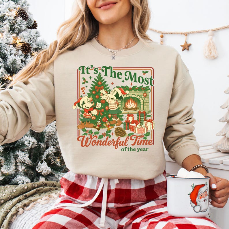 May include: Beige sweatshirt with a Christmas-themed graphic. The design features Mickey and Minnie Mouse, a Christmas tree, a fireplace, and the text "It's The Most Wonderful Time of the Year".