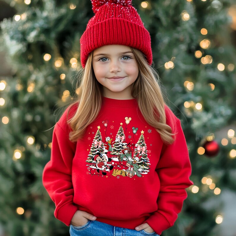 May include: A red Christmas sweater with a festive design featuring Mickey Mouse, Minnie Mouse, Donald Duck, and Goofy. The characters are standing in front of Christmas trees with presents and snowflakes.