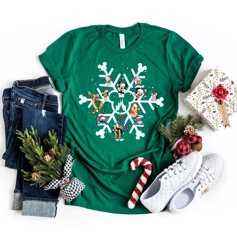 May include: A green t-shirt with a white snowflake design featuring various Disney characters wearing Santa hats. The characters include Mickey Mouse, Donald Duck, Goofy, Pluto, and others. The snowflake design also includes the iconic Disney castle.