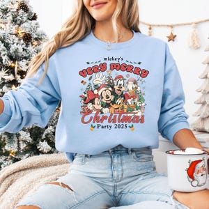 Puede incluir: Sudadera azul claro con un dise&ntilde;o festivo de personajes de Disney, incluyendo Mickey y Minnie Mouse, celebrando la Navidad. El texto "mickey's Very Merry Christmas Party 2025" est&aacute; impreso en rojo y verde sobre los personajes.