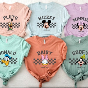 May include: Six pastel-colored t-shirts featuring Disney characters Pluto, Mickey, Minnie, Donald, Daisy, and Goofy. Each shirt displays the character's name above a checkered stripe and the year they debuted. The shirts are a soft, comfortable material.