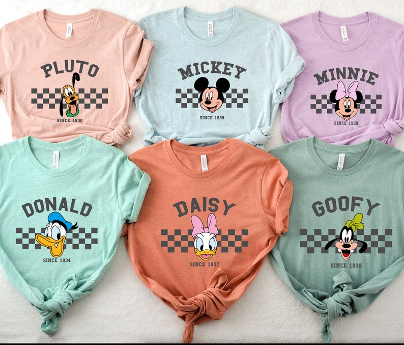 May include: Six pastel-colored t-shirts featuring Disney characters Pluto, Mickey, Minnie, Donald, Daisy, and Goofy. Each shirt displays the character's name above a checkered stripe and the year they debuted. The shirts are a soft, comfortable material.
