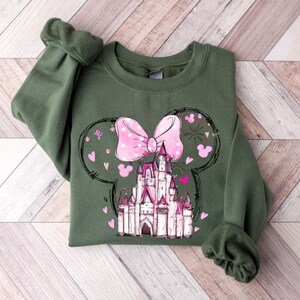 May include: A green sweatshirt with a pink bow and a white castle graphic. The castle is surrounded by pink hearts and stars. The graphic is in the shape of Mickey Mouse ears.