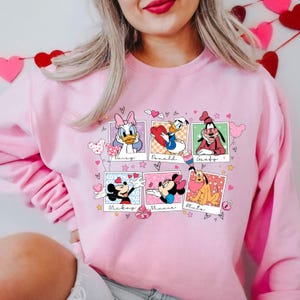 K&ouml;nnte beinhalten: Rosafarbener Pullover mit einem Grafikdruck von Mickey Mouse, Minnie Mouse, Donald Duck, Daisy Duck, Goofy und Pluto in Polaroid-Rahmen mit Herzen und Sternen. Der Text "Mickey", "Minnie", "Donald", "Daisy", "Goofy" und "Pluto" ist unter jedem Rahmen gedruckt.