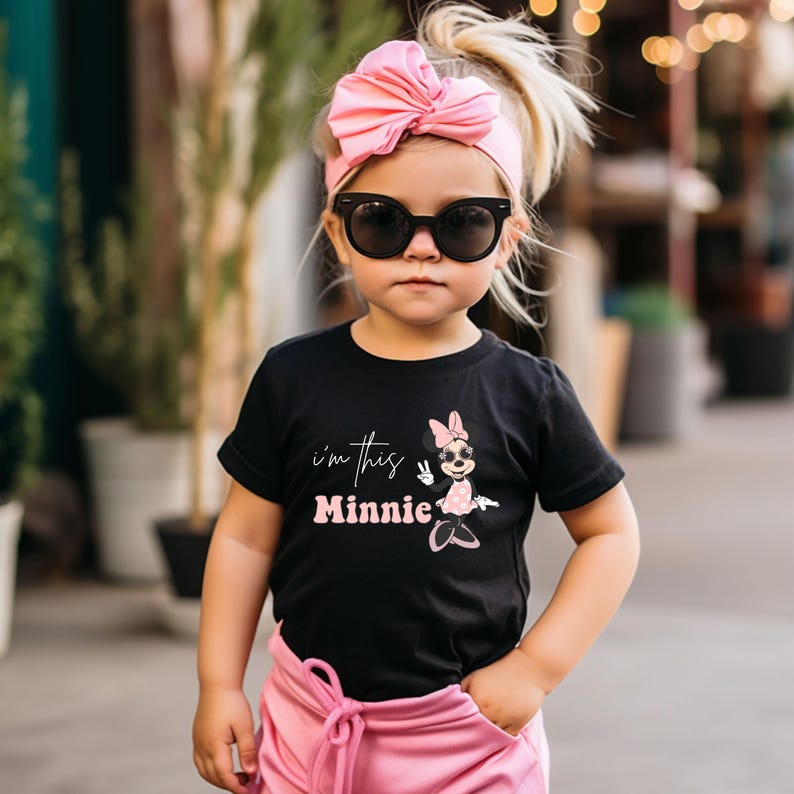 Puede incluir: Una camiseta negra con un gr&aacute;fico rosa de Minnie Mouse y el texto "i'm this Minnie" en la parte delantera. La camiseta la lleva puesta una ni&ntilde;a con gafas de sol rosas y una diadema rosa con lazo.