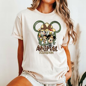 May include: A white t-shirt with a green Mickey Mouse ears design featuring Disney characters from Animal Kingdom. The text "ANIMAL KINGDOM" is printed below the design.