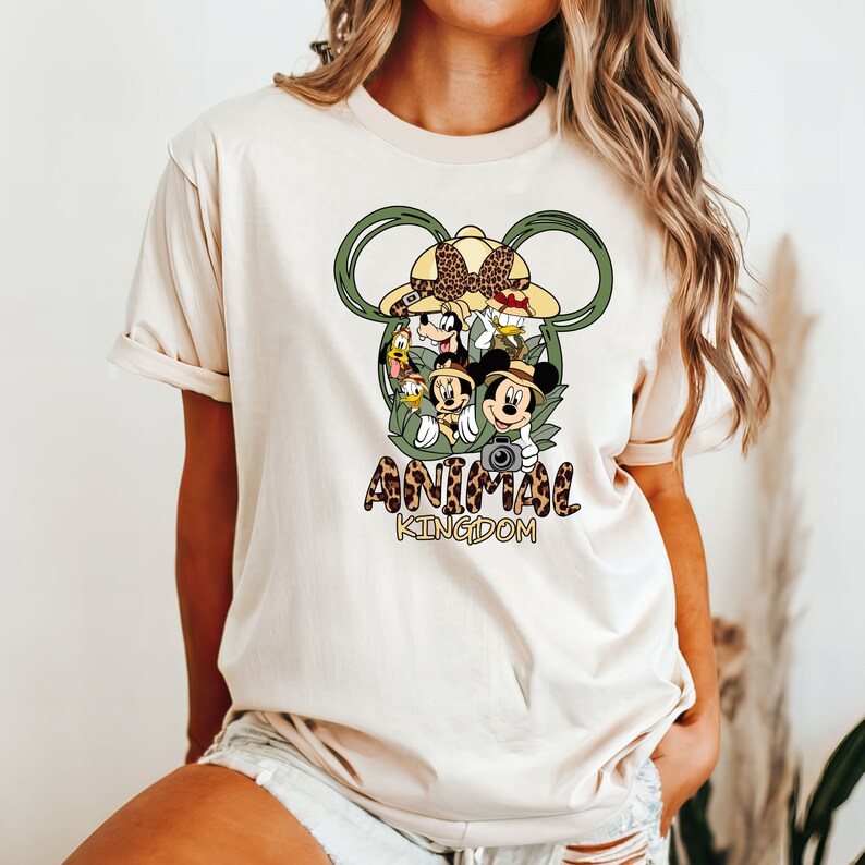 May include: A white t-shirt with a green Mickey Mouse ears design featuring Disney characters from Animal Kingdom. The text "ANIMAL KINGDOM" is printed below the design.
