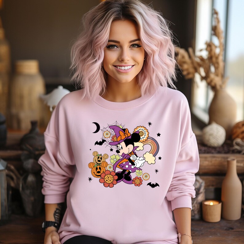 K&ouml;nnte beinhalten: Hellrosa Sweatshirt mit einem Halloween-Motiv von Minnie Mouse. Das Design zeigt Minnie in einem Hexenkost&uuml;m, K&uuml;rbisse, Blumen, einen Regenbogen, Flederm&auml;use und einen Mond. Das Sweatshirt hat lange &Auml;rmel und einen Rundhalsausschnitt.