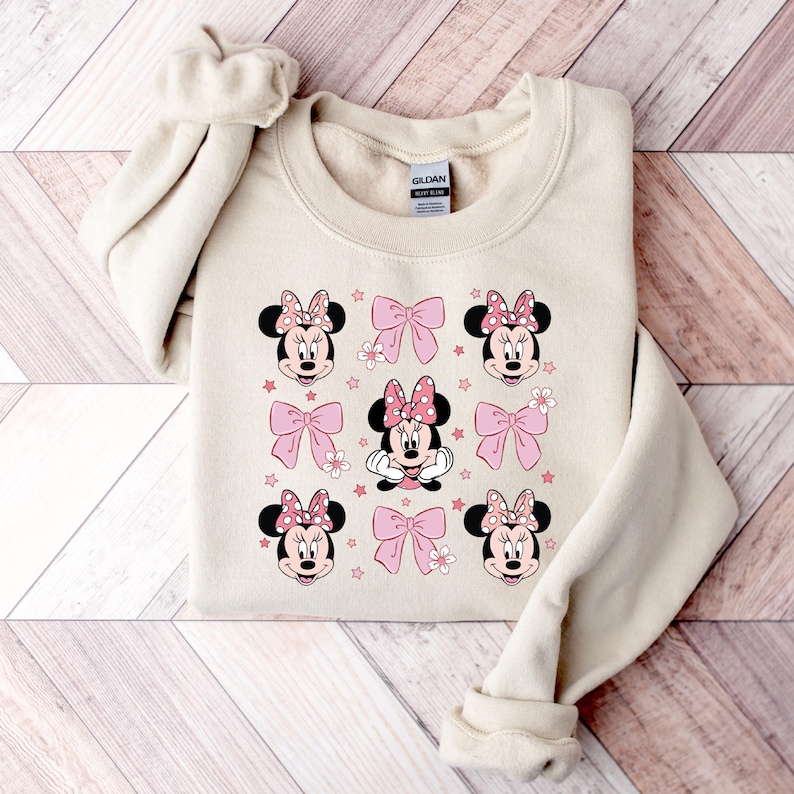 May include: A beige sweatshirt with a repeating pattern of pink bows and cartoon Minnie Mouse faces. The sweatshirt has a Gildan brand tag.