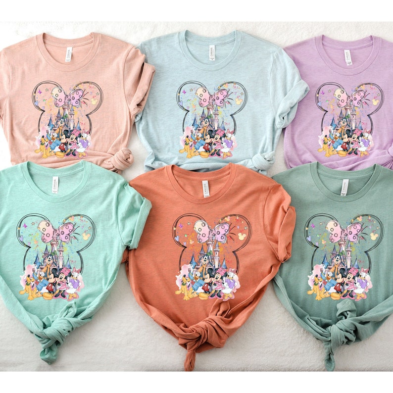 May include: Five t-shirts with a colorful graphic of Mickey Mouse and friends in front of Cinderella's Castle. The shirts are in various colors, including pink, light blue, lavender, green, and orange.