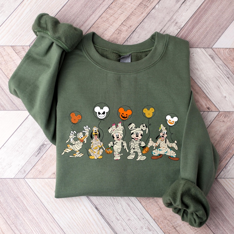 Peut inclure: Un sweat-shirt vert avec un imprim&eacute; de Mickey Mouse, Minnie Mouse, Donald Duck, Dingo et Pluto habill&eacute;s en momies tenant des ballons. Les ballons sont en forme de t&ecirc;te de Mickey Mouse et ont des couleurs d'Halloween.