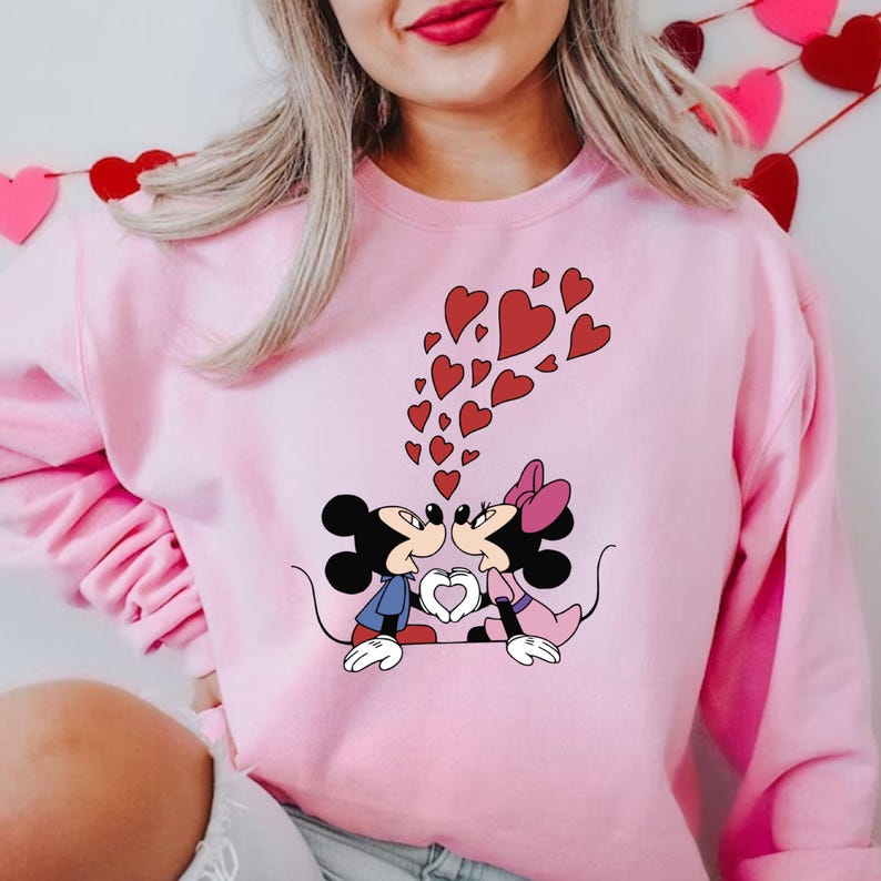 May include: Pink sweatshirt with a graphic of Mickey Mouse and Minnie Mouse kissing, surrounded by red hearts.