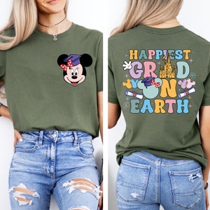 Puede incluir: Una camiseta verde con un gr&aacute;fico de Mickey Mouse con un gorro de graduaci&oacute;n y el texto "Happiest Grad on Earth" en la parte posterior. La camiseta est&aacute; siendo modelada por una persona que lleva pantalones vaqueros azules.