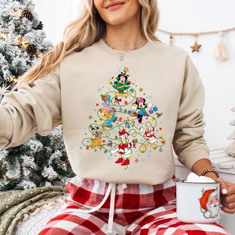 K&ouml;nnte beinhalten: Beiges Sweatshirt mit einem Weihnachtsbaum-Design aus Disney-Figuren wie Mickey und Minnie Mouse. Der Baum ist mit Kugeln und Lichterketten geschm&uuml;ckt. Das Sweatshirt wird mit einer rot-wei&szlig; karierten Pyjamahose getragen.