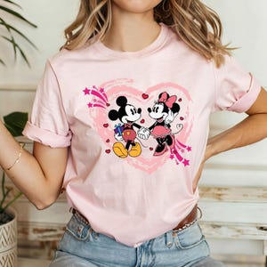 May include: A light pink t-shirt featuring a graphic of Mickey and Minnie Mouse holding hands inside a pink heart. Mickey Mouse is holding a gift. The design includes pink stars and hearts.
