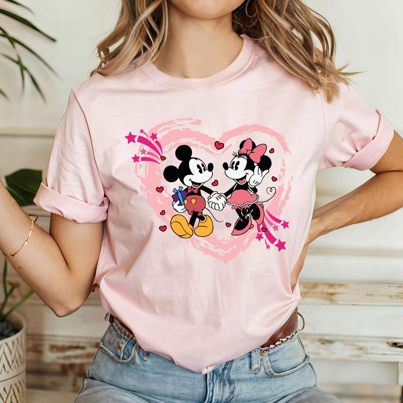 May include: A light pink t-shirt featuring a graphic of Mickey and Minnie Mouse holding hands inside a pink heart. Mickey Mouse is holding a gift. The design includes pink stars and hearts.
