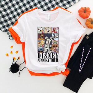 Pu&ograve; includere: T-shirt bianco con bordi arancioni con una grafica Disney Spooky Tour. La grafica include vari personaggi Disney in costume di Halloween. Anche pantaloni neri e zucche decorative sono nell'immagine.