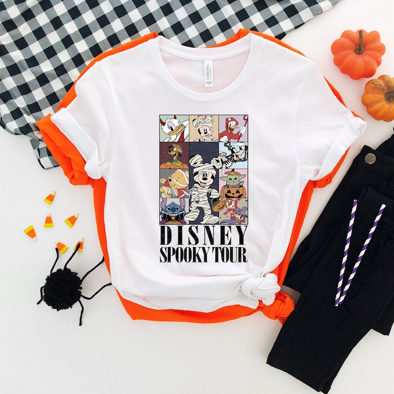 Pu&ograve; includere: T-shirt bianco con bordi arancioni con una grafica Disney Spooky Tour. La grafica include vari personaggi Disney in costume di Halloween. Anche pantaloni neri e zucche decorative sono nell'immagine.