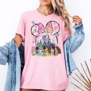 May include: Pink t-shirt with a graphic of Cinderella's castle, a pink bow, and seven princesses in front of the castle.