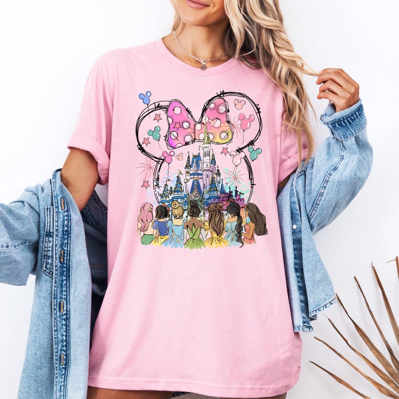 May include: Pink t-shirt with a graphic of Cinderella's castle, a pink bow, and seven princesses in front of the castle.
