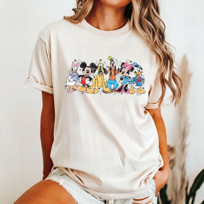 Puede incluir: Una camiseta de color crema con un estampado gr&aacute;fico de Mickey Mouse, Minnie Mouse, Donald Duck, Goofy y Pluto. Los personajes est&aacute;n todos sonriendo y de pie juntos.