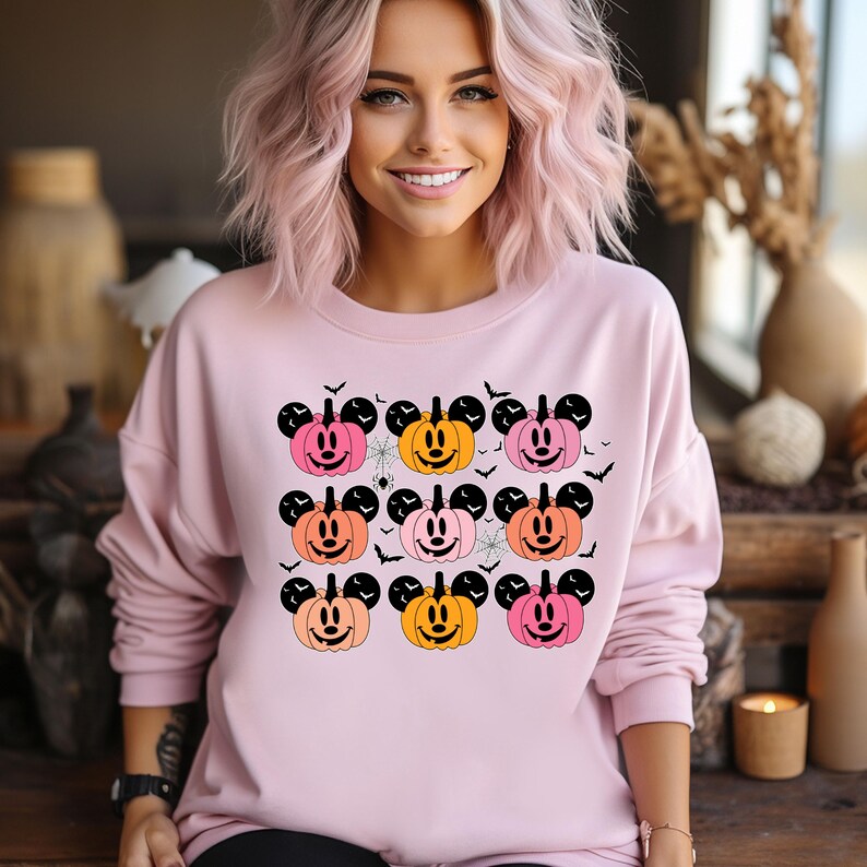 May include: A pink sweatshirt with a Halloween theme featuring nine orange, pink, and yellow pumpkins with Mickey Mouse ears. The pumpkins have black eyes and a black smile.