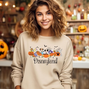 K&ouml;nnte beinhalten: Beiges Sweatshirt mit Rundhalsausschnitt und einem Halloween-Disneyland-Motiv. Die Grafik zeigt K&uuml;rbisse, Flederm&auml;use, Spinnen und Disney-Figuren. Das Wort "Disneyland" ist in schwarzer Schrift geschrieben.