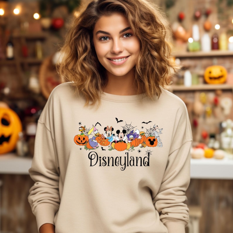 K&ouml;nnte beinhalten: Beiges Sweatshirt mit Rundhalsausschnitt und einem Halloween-Disneyland-Motiv. Die Grafik zeigt K&uuml;rbisse, Flederm&auml;use, Spinnen und Disney-Figuren. Das Wort "Disneyland" ist in schwarzer Schrift geschrieben.