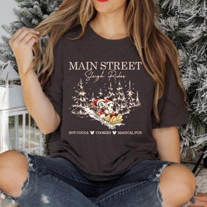 May include: Dark gray t-shirt featuring a winter scene with Mickey and Minnie Mouse on a sleigh. The shirt has the text "Main Street Sleigh Rides" and "Hot Cocoa Cookies Magical Fun" in a festive font. The design includes snow-covered trees.