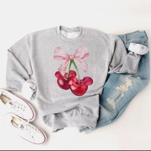 Puede incluir: Una sudadera gris con un lazo rosa y dos cerezas rojas en la parte delantera. Las cerezas tienen tallos y hojas verdes.