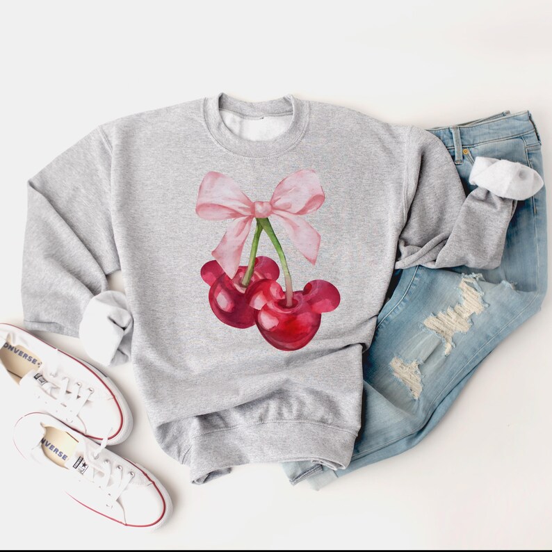 Puede incluir: Una sudadera gris con un lazo rosa y dos cerezas rojas en la parte delantera. Las cerezas tienen tallos y hojas verdes.