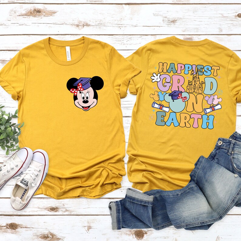 Puede incluir: Dos camisetas amarillas con un tema de graduaci&oacute;n. Una camiseta presenta una imagen de dibujos animados de Mickey Mouse con un gorro de graduaci&oacute;n. La otra camiseta tiene el texto "Happiest Grad on Earth" con una imagen de dibujos animados del castillo de Cenicienta.