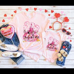 May include: Pink t-shirts with a graphic of Mickey Mouse and friends in front of Cinderella's Castle with the word "Valentine" in pink letters. The shirts are on a wooden background with hearts and other Valentine's Day decorations.