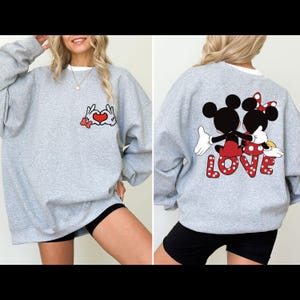 May include: A gray sweatshirt with a Mickey Mouse and Minnie Mouse design on the back. The design features the characters holding hands and the word "LOVE" in white polka dots. The front of the sweatshirt features a design of Mickey Mouse and Minnie Mouse hands forming a heart.