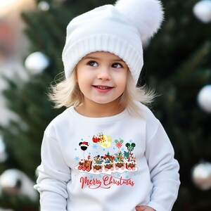 May include: A white long-sleeved sweatshirt with a Christmas-themed design featuring a train with various Disney characters. The train is decorated with snowflakes, candy canes, and a banner that reads "Merry Christmas". The sweatshirt is worn by a young child wearing a white knit hat with a pom-pom.