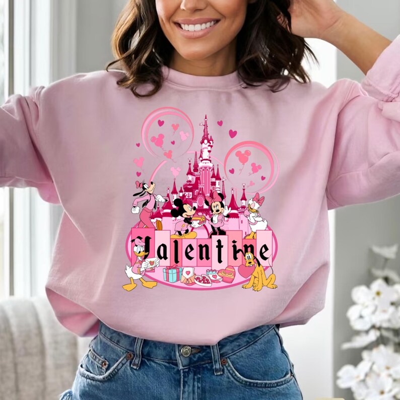 May include: Pink sweatshirt with a Valentine's Day theme featuring Mickey Mouse and friends in front of a pink castle with the word "Valentine" printed on the front.