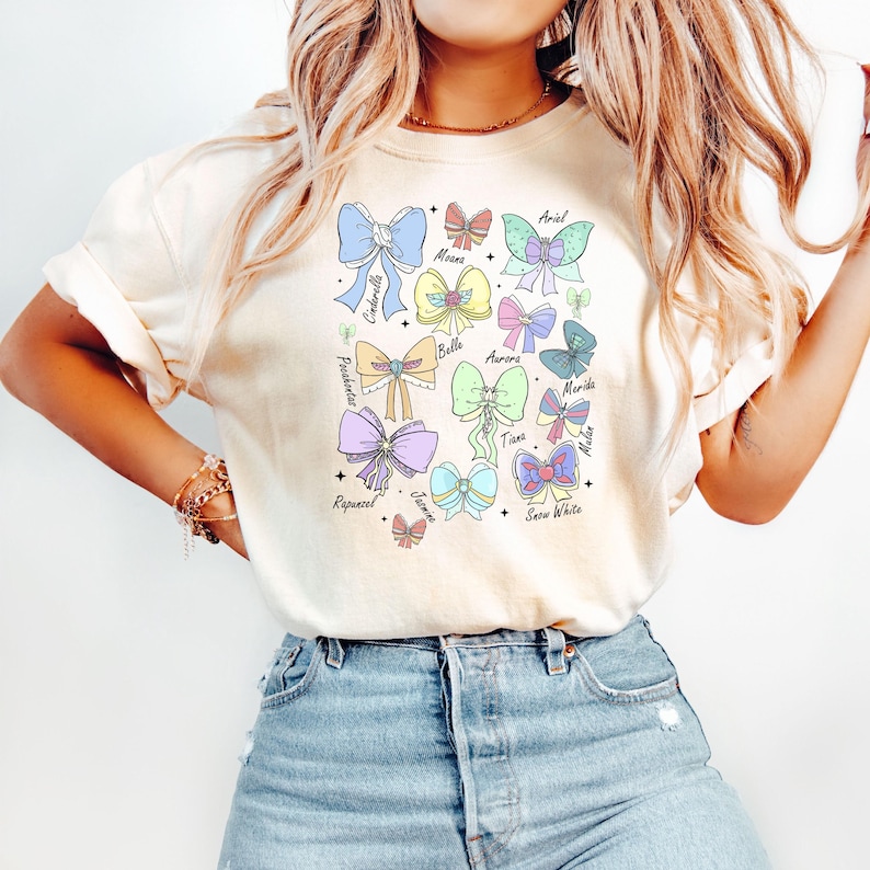 May include: A white t-shirt with a graphic of colorful bows and the names of Disney princesses: Cinderella, Moana, Belle, Aurora, Merida, Rapunzel, Jasmine, Snow White, Ariel, Tiana, and Mulan.