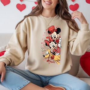 Puede incluir: Sudadera beige con Mickey Mouse, Minnie Mouse y Pluto con atuendo de San Valent&iacute;n. Mickey lleva un sombrero de copa, Minnie un lazo y Pluto tiene cuernos y una correa en forma de coraz&oacute;n. El dise&ntilde;o incluye corazones y rosas.