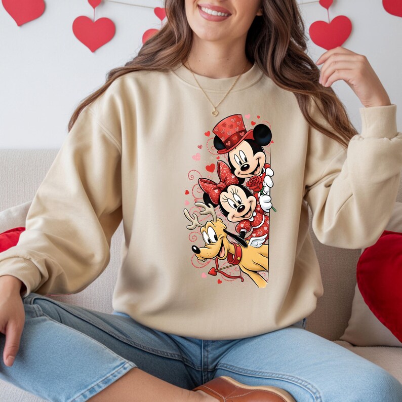 Puede incluir: Sudadera beige con Mickey Mouse, Minnie Mouse y Pluto con atuendo de San Valent&iacute;n. Mickey lleva un sombrero de copa, Minnie un lazo y Pluto tiene cuernos y una correa en forma de coraz&oacute;n. El dise&ntilde;o incluye corazones y rosas.