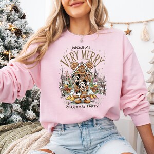 Puede incluir: Sudadera rosa claro con un dise&ntilde;o festivo de Mickey Mouse y sus amigos. El dise&ntilde;o incluye una casa de jengibre y el texto "Mickey's Very Merry Christmas Party". La sudadera es de cuello redondo y manga larga.