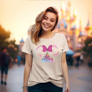 May include: A light beige t-shirt with a pink bow and a glitter castle graphic. The castle is a silhouette of Cinderella's castle.