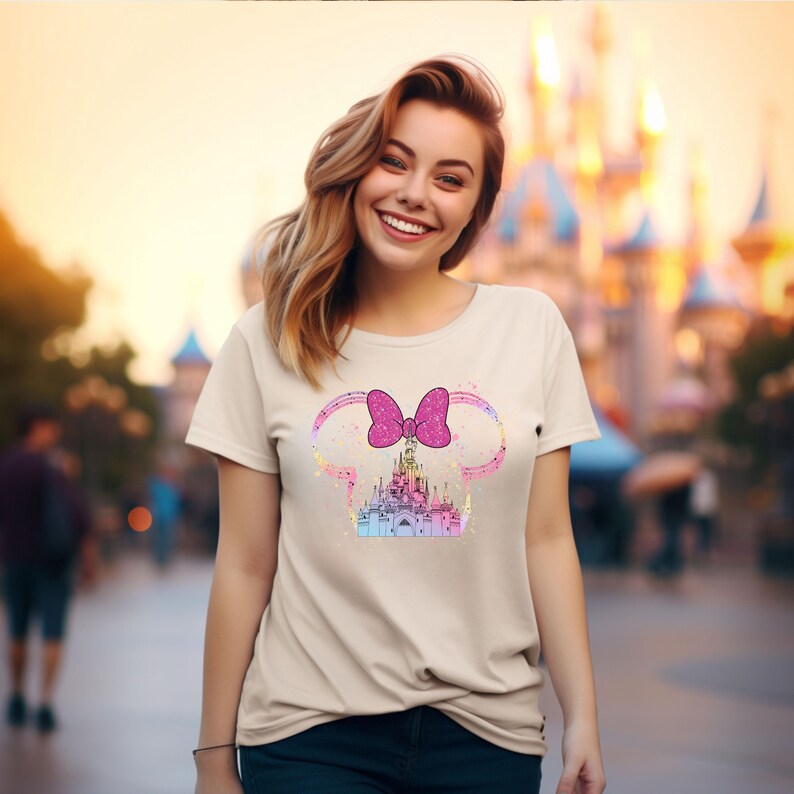 May include: A light beige t-shirt with a pink bow and a glitter castle graphic. The castle is a silhouette of Cinderella's castle.
