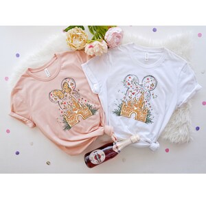 May include: Two pink and white t-shirts with a graphic of a gingerbread castle with Mickey Mouse ears on top. The castle is surrounded by fireworks and confetti. The shirts are laid out on a white surface with a bottle of sparkling wine.