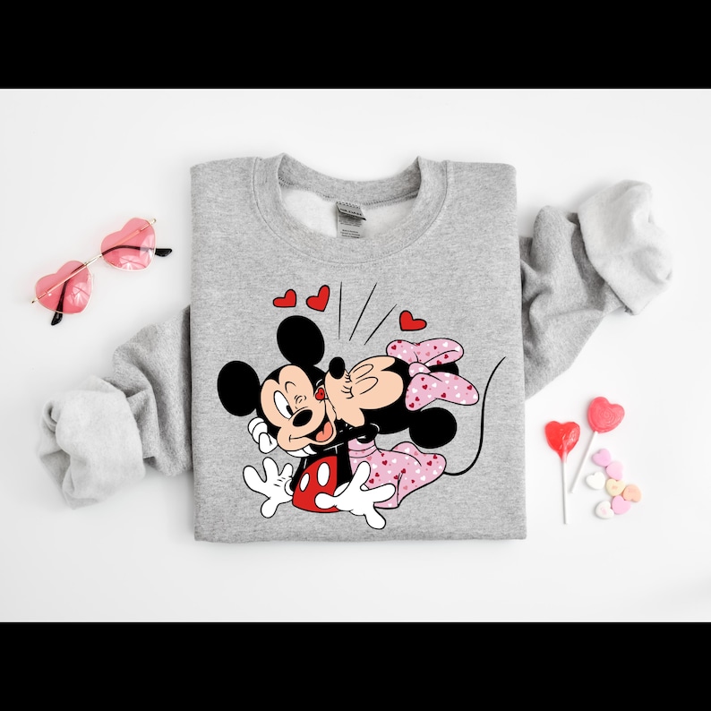 May include: A grey sweatshirt featuring a printed design of Mickey Mouse and Minnie Mouse kissing. The design is surrounded by red hearts.