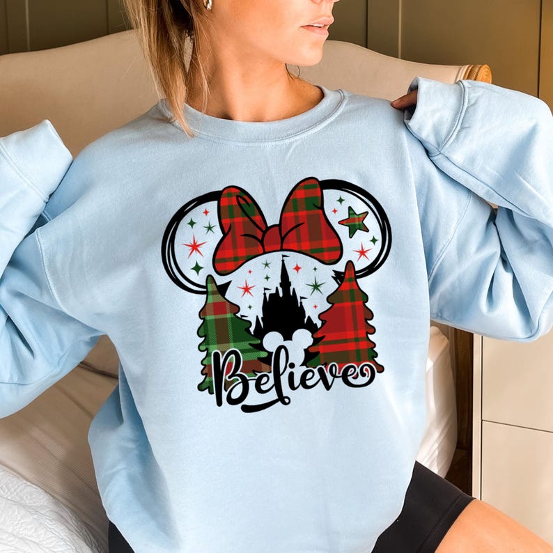 Puede incluir: Sudadera azul claro con un dise&ntilde;o de orejas de Minnie Mouse en cuadros rojos y verdes, una silueta del castillo de Cenicienta y la palabra "Believe".
