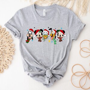 May include: A gray t-shirt with a Christmas-themed design featuring Mickey Mouse, Minnie Mouse, Donald Duck, Goofy, and Pluto wearing Santa hats and scarves.